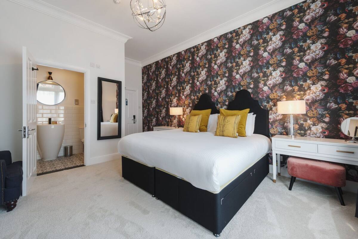 A bedroom with a large bed, a dresser, a mirror, and a floral wallpaper.