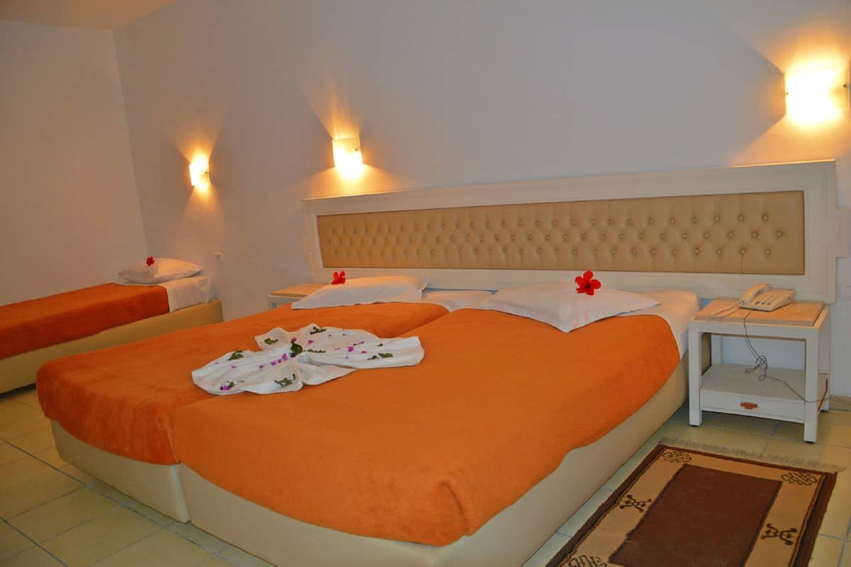 Habitación de hotel con dos camas, cada una con ropa de cama naranja y almohadas blancas. Hay dos mesitas de noche con lámparas y una alfombra pequeña en el suelo.
