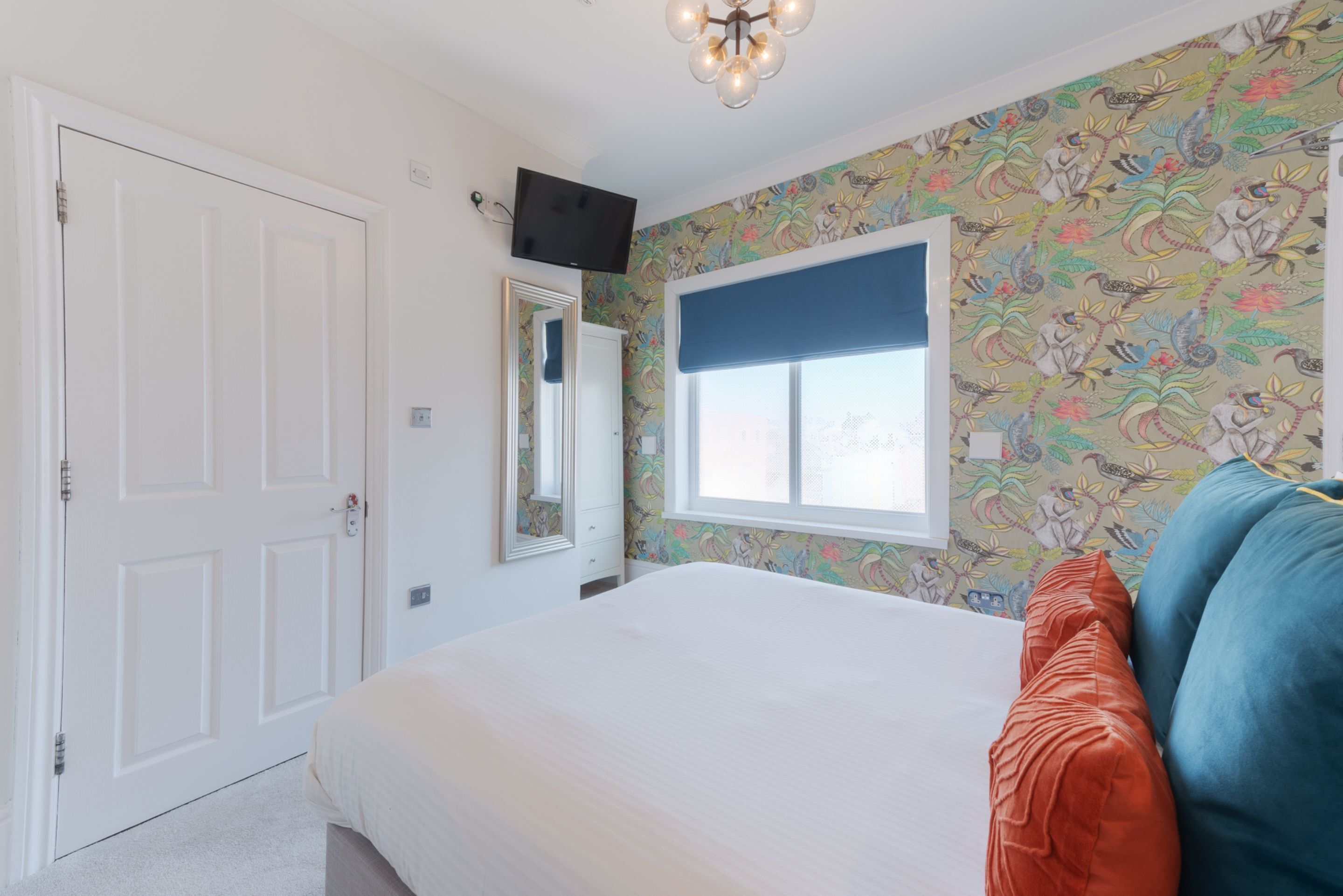A bedroom with a bed, a window with a blue blind, a door, and a wall with a floral pattern.