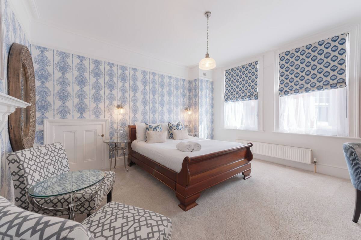 A bedroom with a wooden bed, patterned wallpaper, a glass table, and a chair.