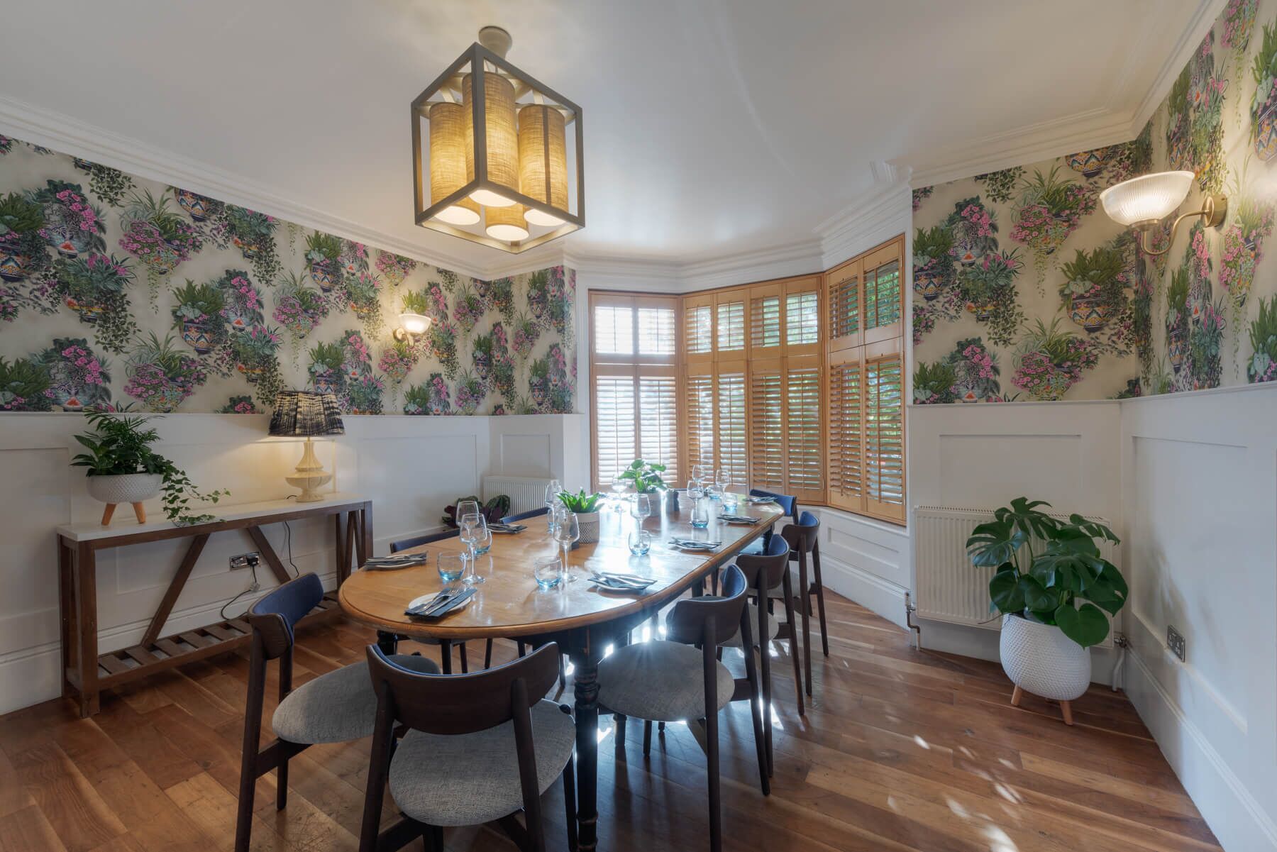 Un comedor con mesa de madera dispuesta para seis personas, rodeada de sillas. Hay un ventanal con contraventanas de madera, un radiador y una planta en maceta sobre el suelo.