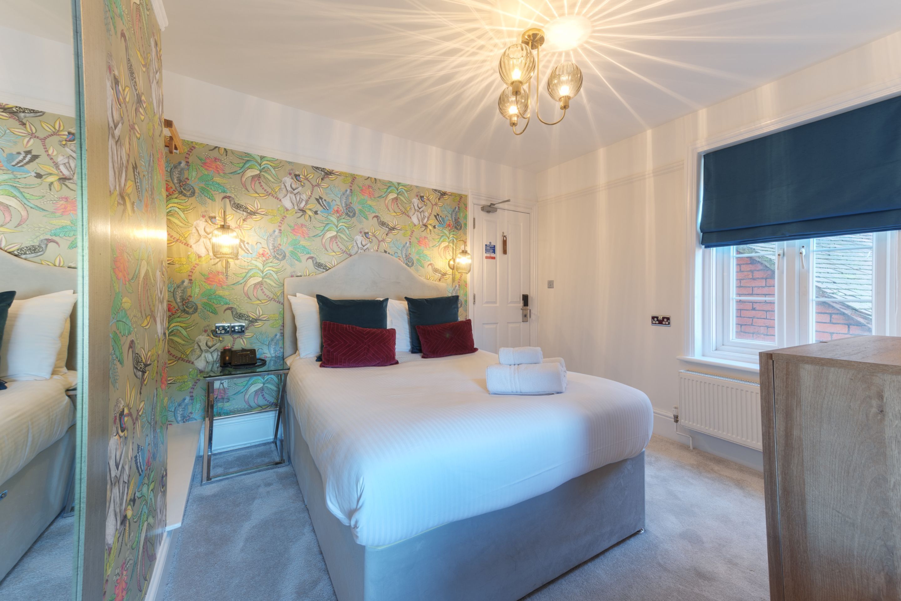 A bedroom with a large bed, a bedside table, a mirror, a wall with floral wallpaper, a ceiling light fixture, and a window with a blind.