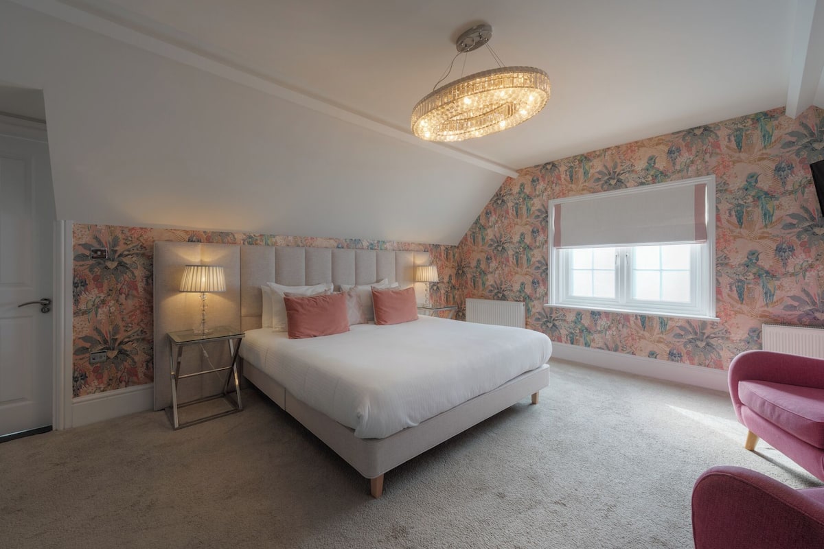 A bedroom with a large bed, a chandelier, a window, and floral wallpaper.