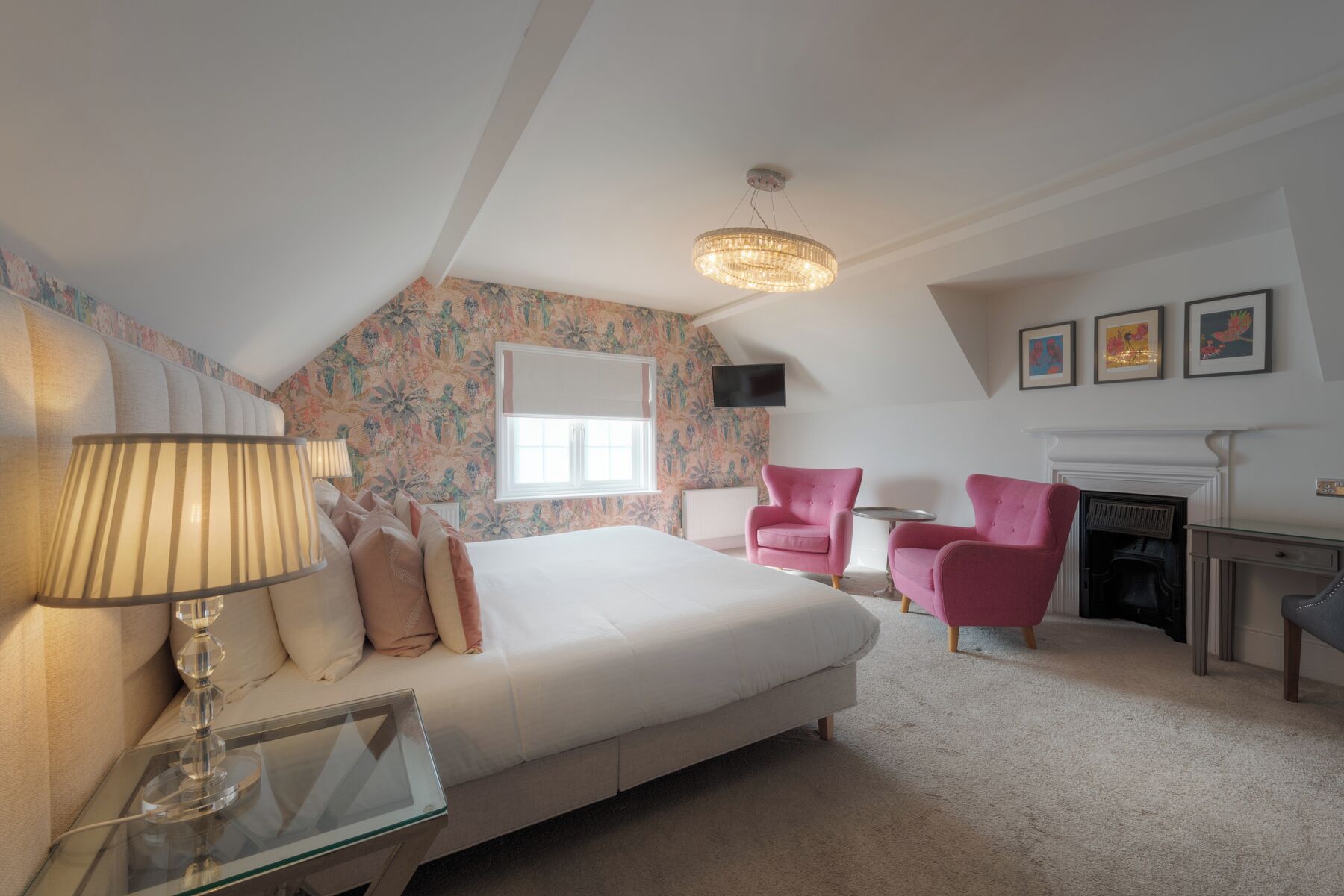 Een slaapkamer met een groot bed, twee roze fauteuils, een open haard, een televisie aan de muur en een raam met uitzicht.