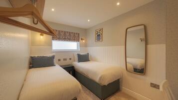 A bedroom with two single beds, a window with a floral curtain, a wall-mounted mirror, and a ceiling fan.