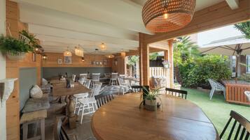 Un restaurant chaleureux avec des tables et des chaises en bois, des plantes suspendues et un grand luminaire tressé.