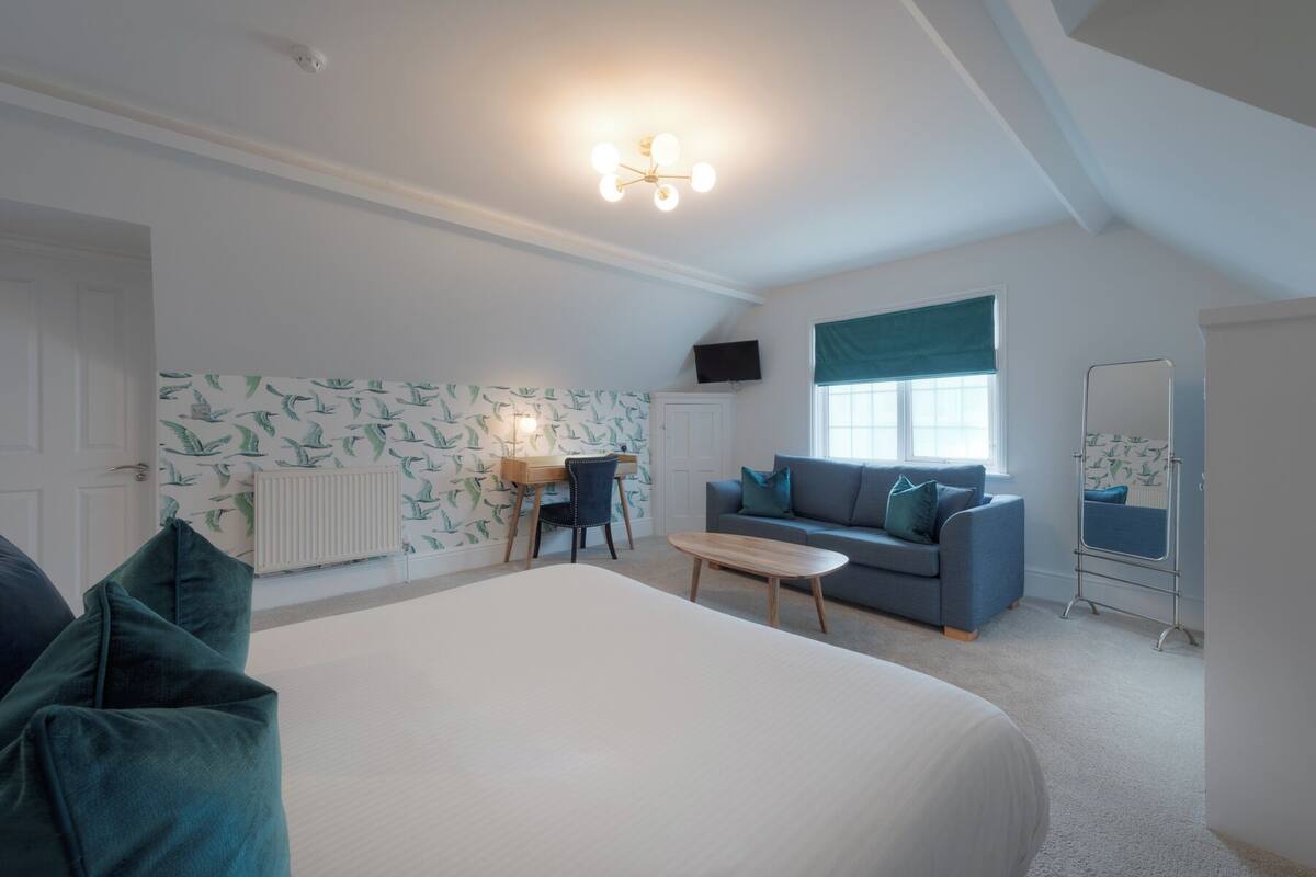 A bedroom with a large bed, a desk with a chair, a sofa, a TV mounted on the wall, and a radiator.