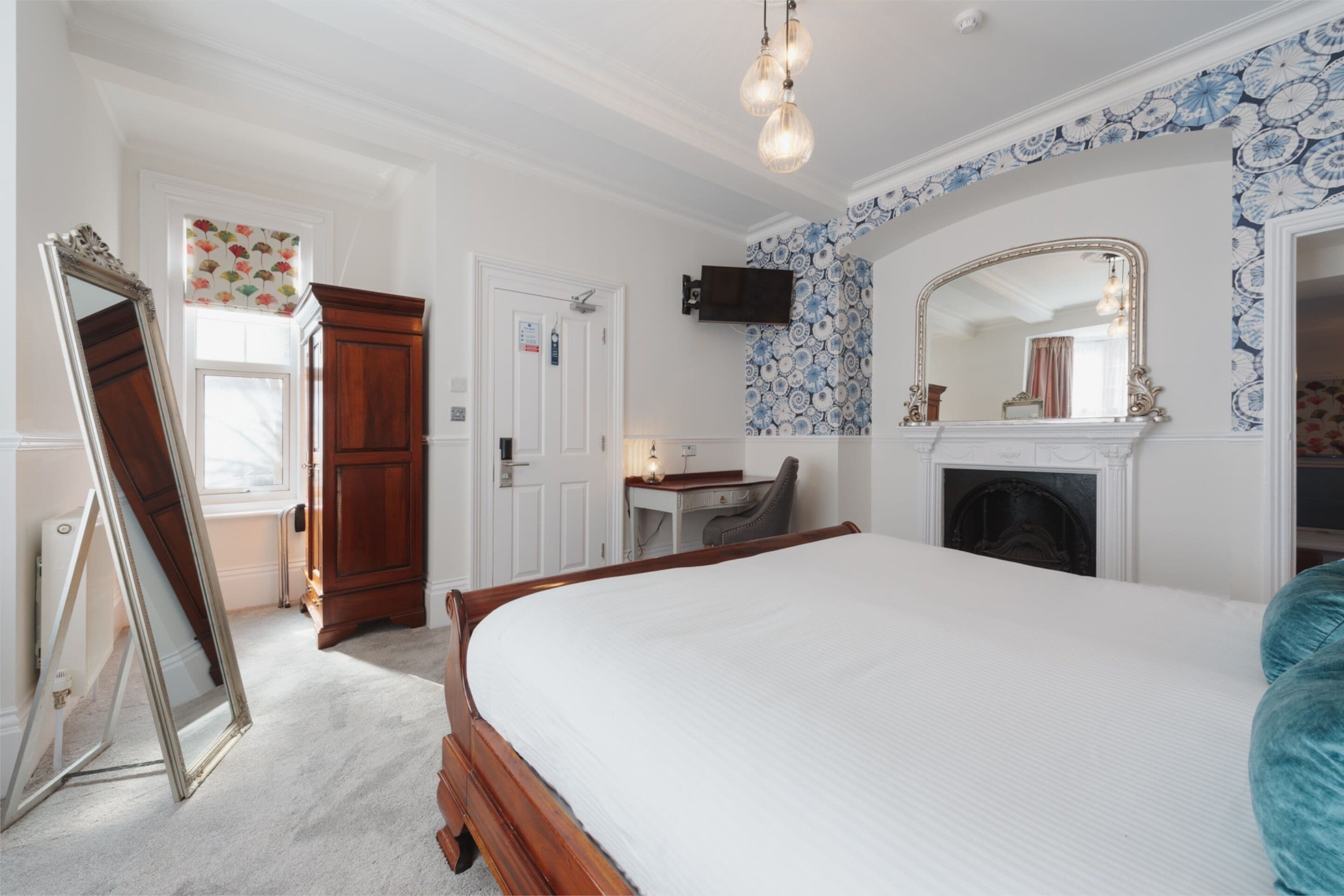A bedroom with a large bed, a wooden wardrobe, a desk, a chair, a television, a fireplace, and a mirror.