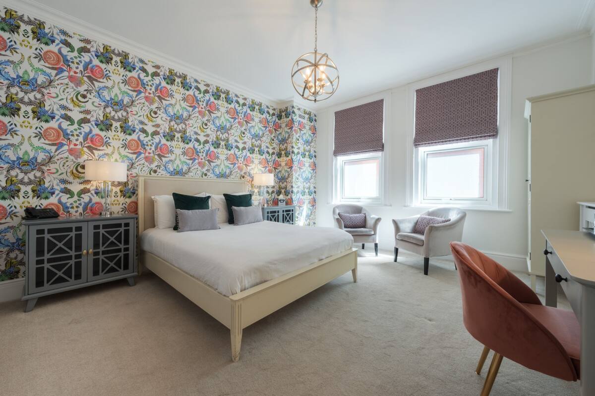 A bedroom with a bed, two chairs, a desk, a lamp, a wardrobe, and a patterned wallpaper.