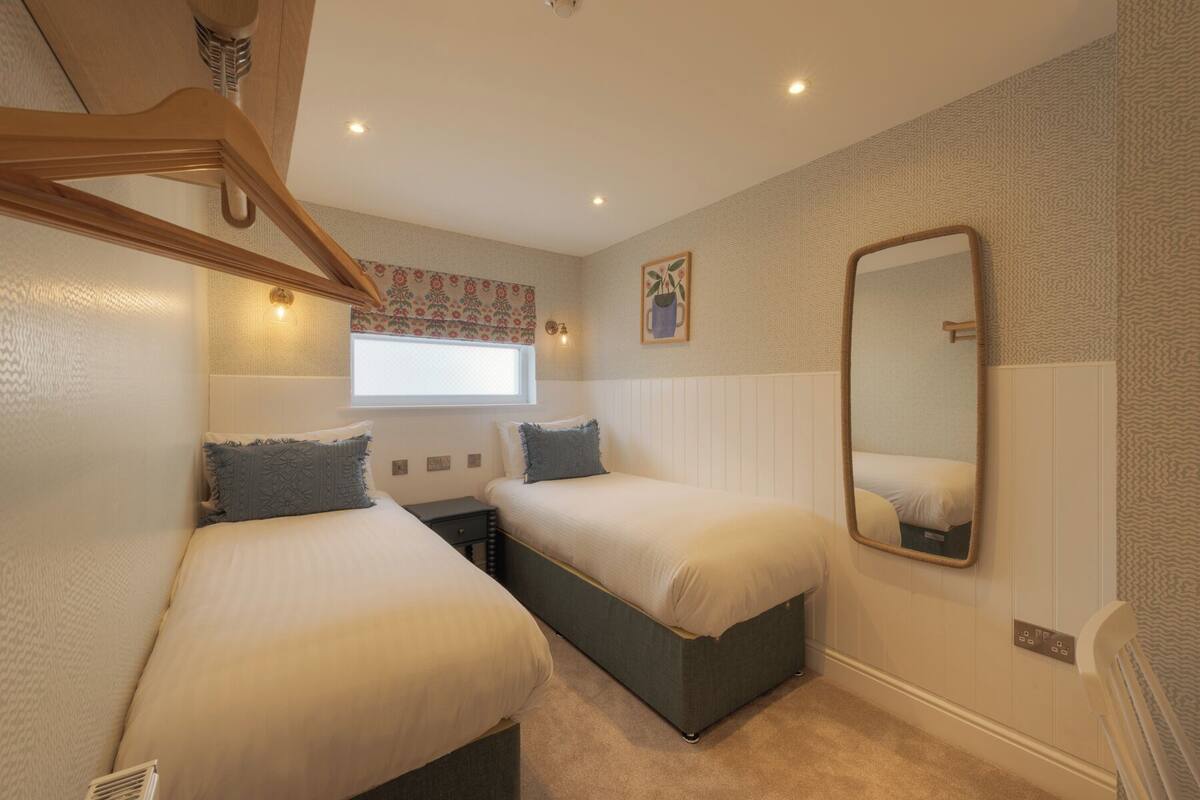 A bedroom with two beds, a window with a floral curtain, a wall-mounted mirror, and a ceiling fan.
