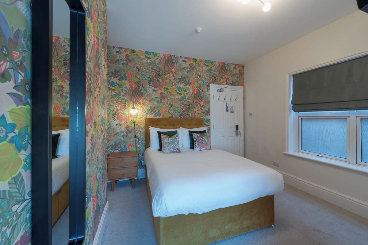 A bedroom with a floral wallpaper, a bed with a wooden headboard, a bedside table, a wardrobe, and a window with a blind.