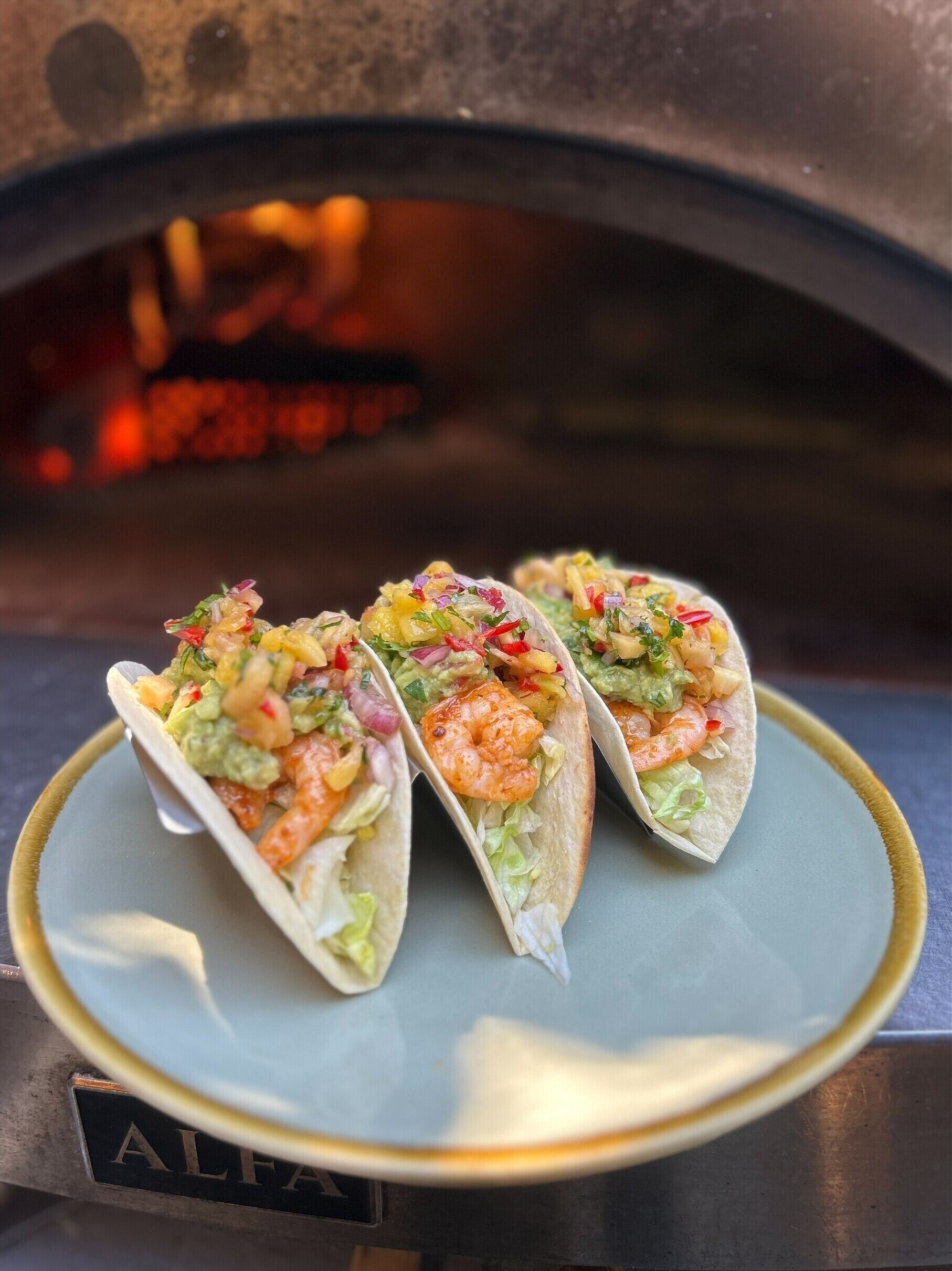 Three tacos on a plate with a pizza oven in the background.
