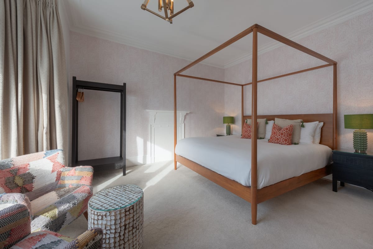 A four-poster bed with a white bedspread and multiple pillows, a round coffee table with a patterned top, a patterned armchair, and a small side table with a lamp.