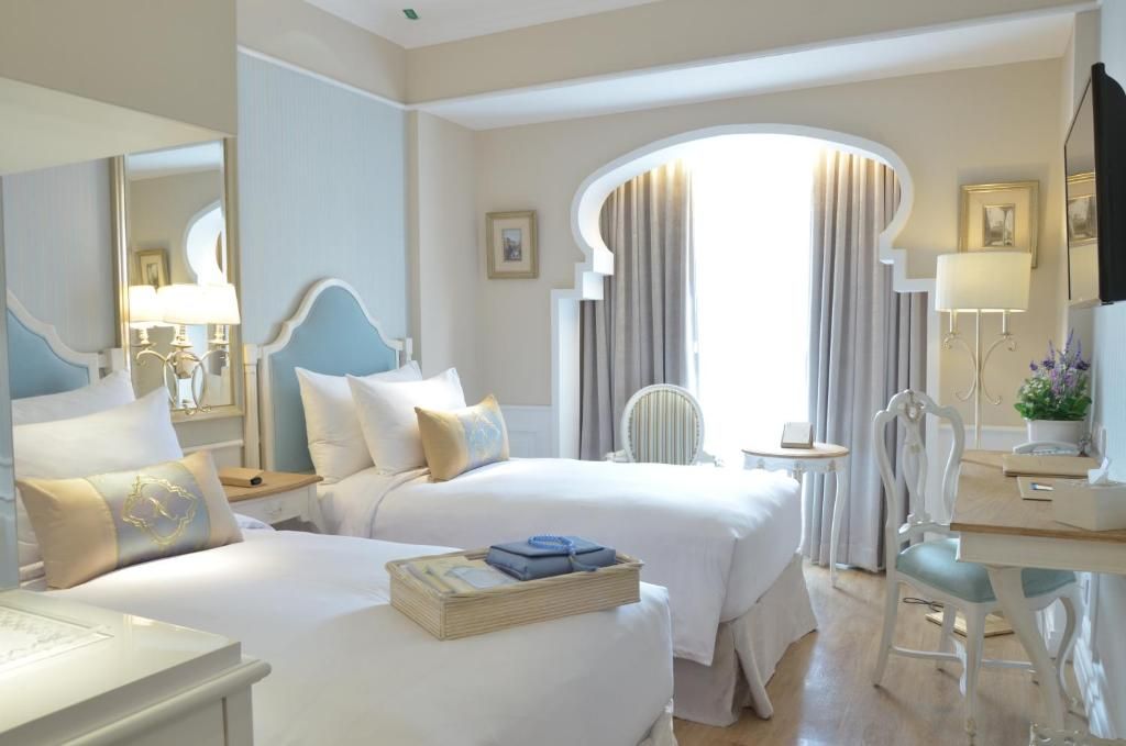 A hotel room with two beds, a desk, a chair, and a large window with curtains.