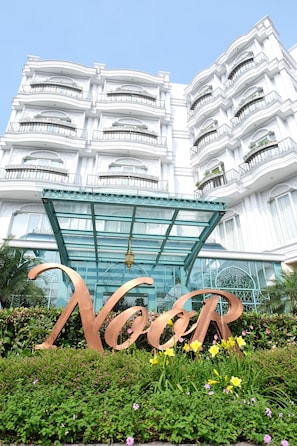 Entrada de un hotel con un gran cartel que dice 'Noor', un toldo de cristal y un edificio blanco con balcones.