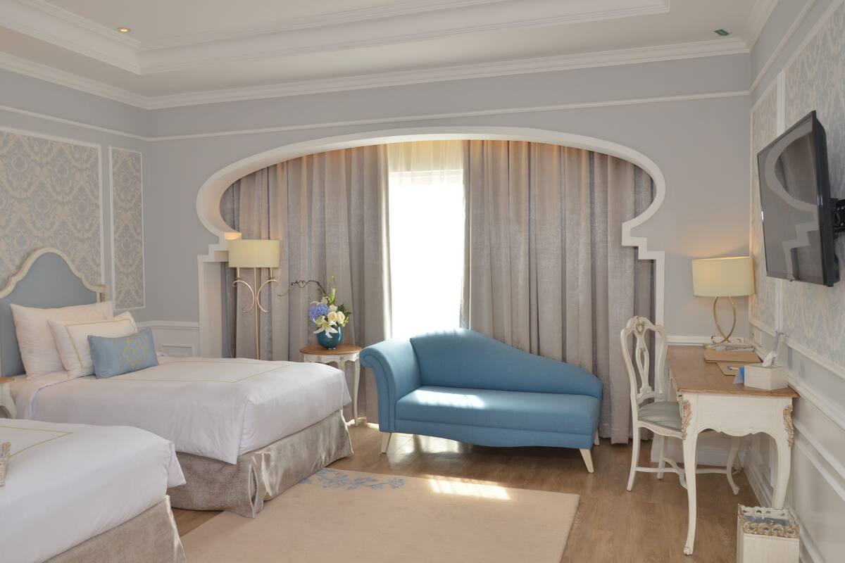 A hotel room with two beds, a blue armchair, a white chair, a TV, and a window with curtains.