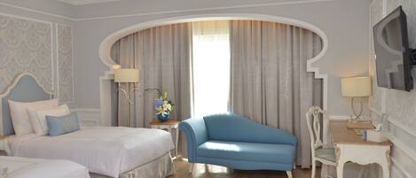 A hotel room with two beds, a blue armchair, a white chair, a TV, and a window with curtains.