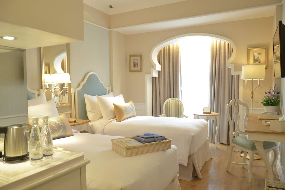 A hotel room with two beds, a desk, a chair, a small table, and a large window with curtains.