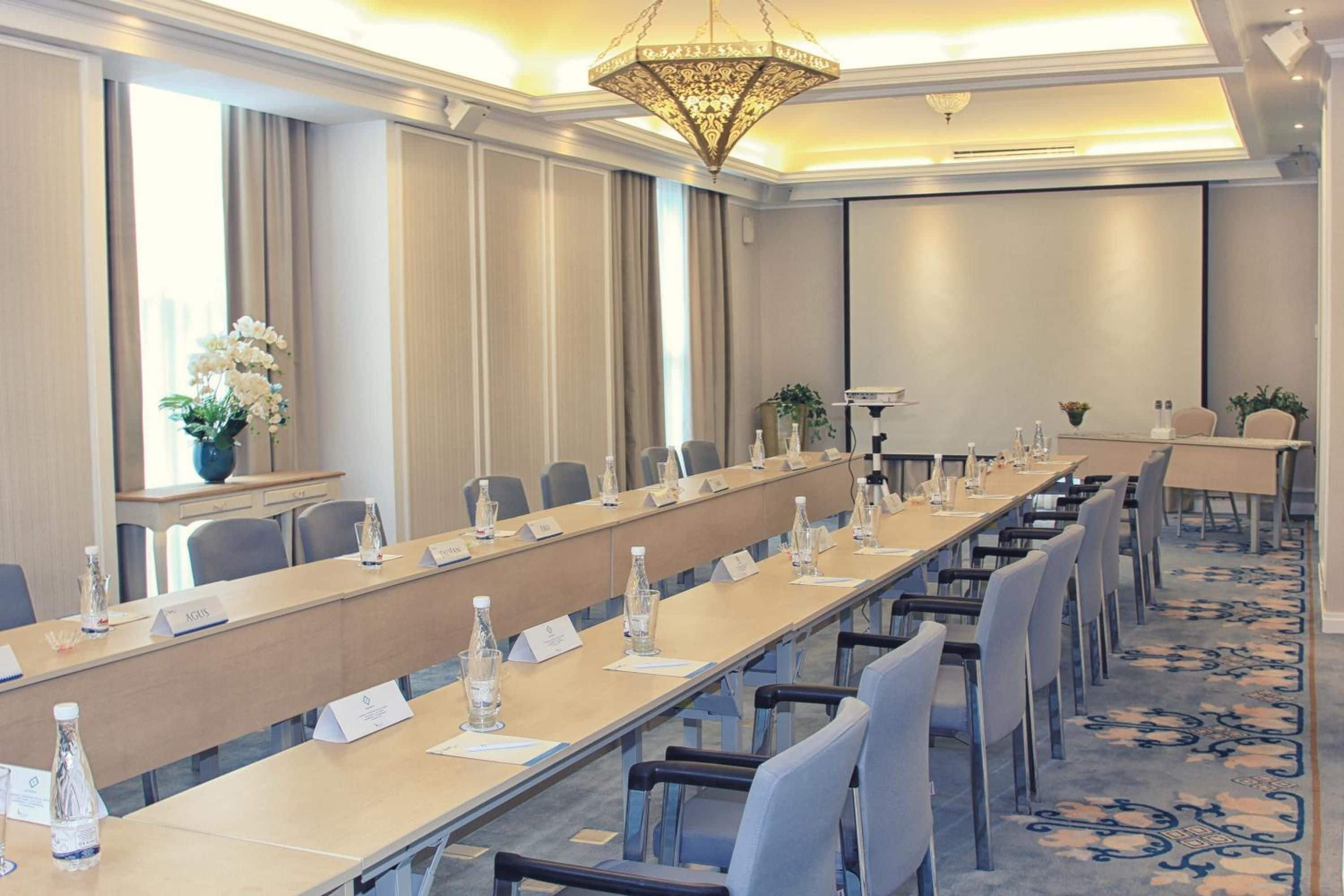 Un salón de conferencias con una larga mesa, sillas, tarjetas de identificación, botellas de agua y una araña de cristal.