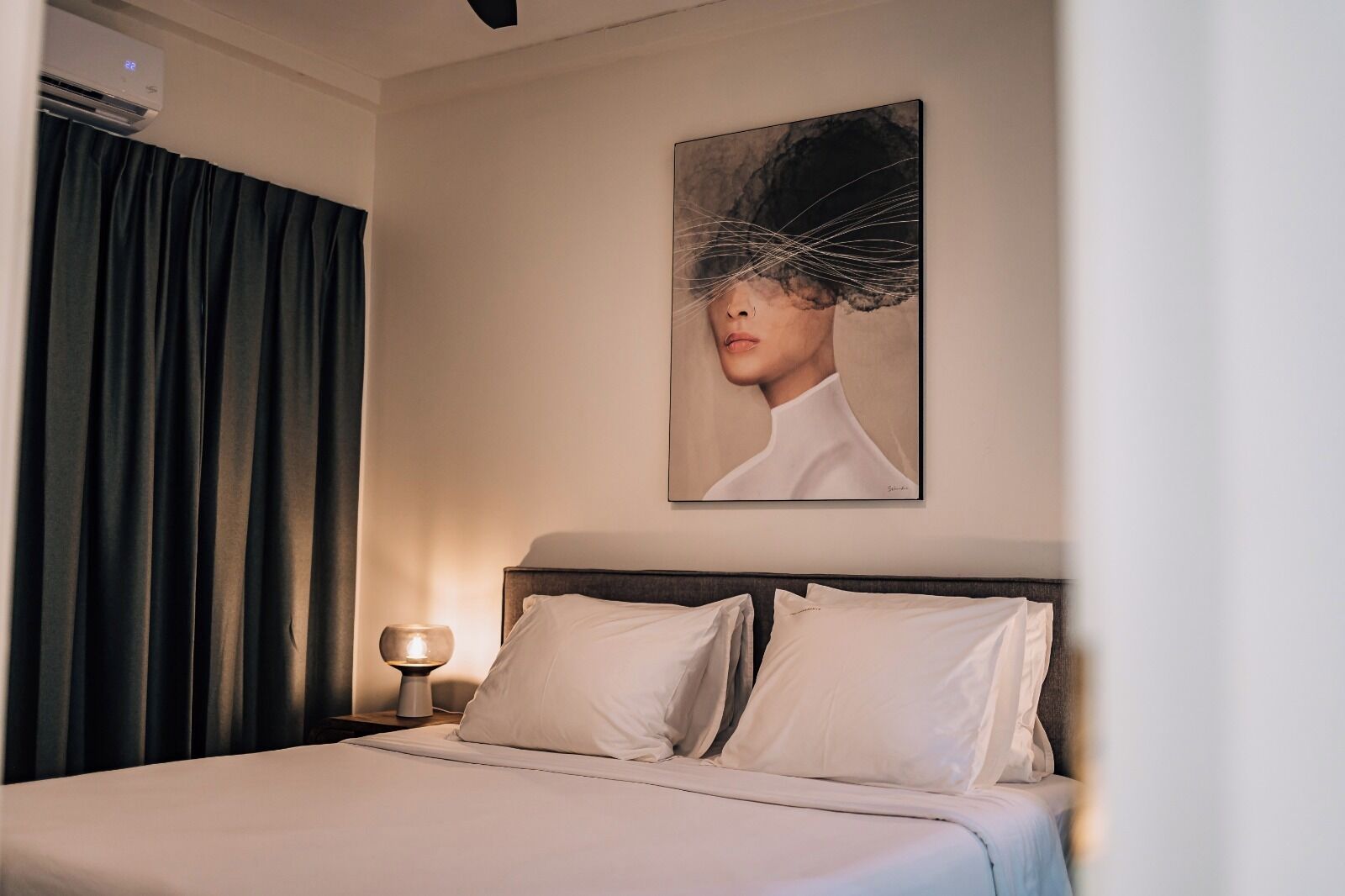 Una habitación de hotel con una cama, una mesita de noche con lámpara, un cuadro en la pared y cortinas oscuras.