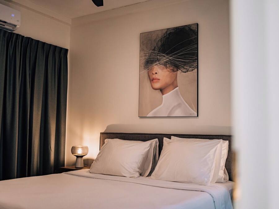 Una habitación de hotel con una cama, una mesita de noche con lámpara, un cuadro en la pared y cortinas oscuras.