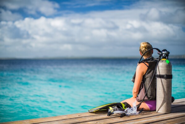 A person sitting on a wooden dock with a diving gear and a scuba tank beside them, overlooking a clear blue sea.