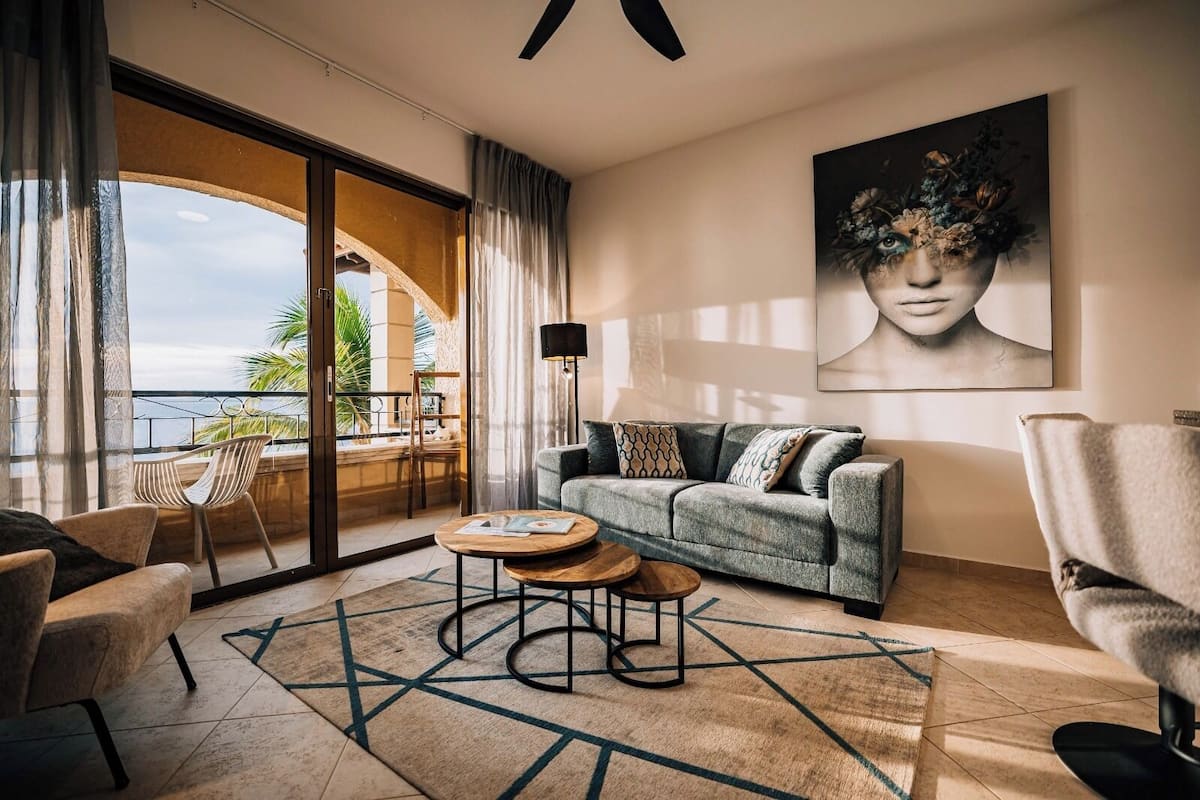A modern living room with a sofa, coffee table, and chairs, a large painting on the wall, and a view of the ocean through a sliding glass door.