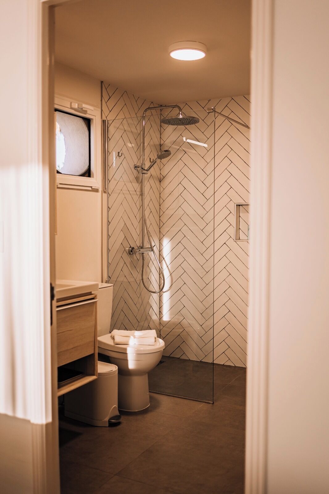 Un baño moderno con un patrón de baldosas en espiga, cabina de ducha de vidrio y un inodoro con tapa blanca.