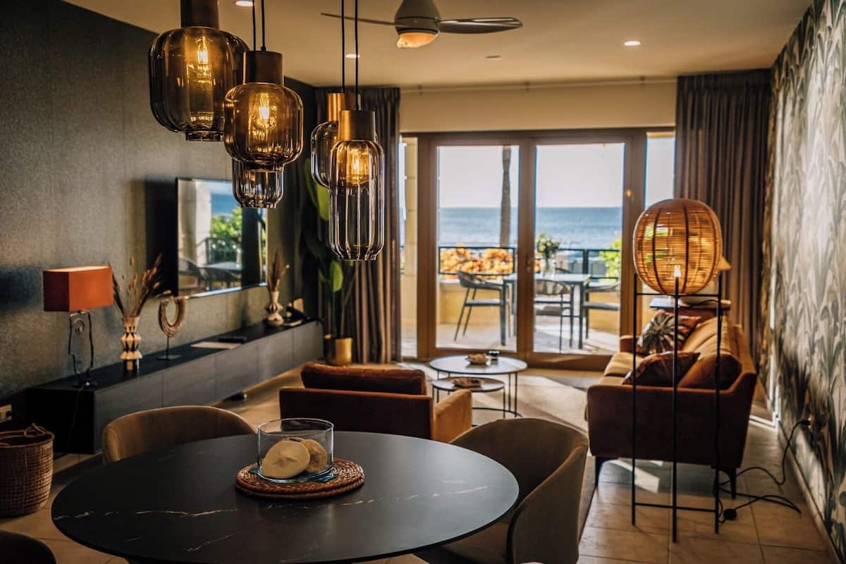 A modern living room with a round table, sofas, and a view of the sea through large windows.