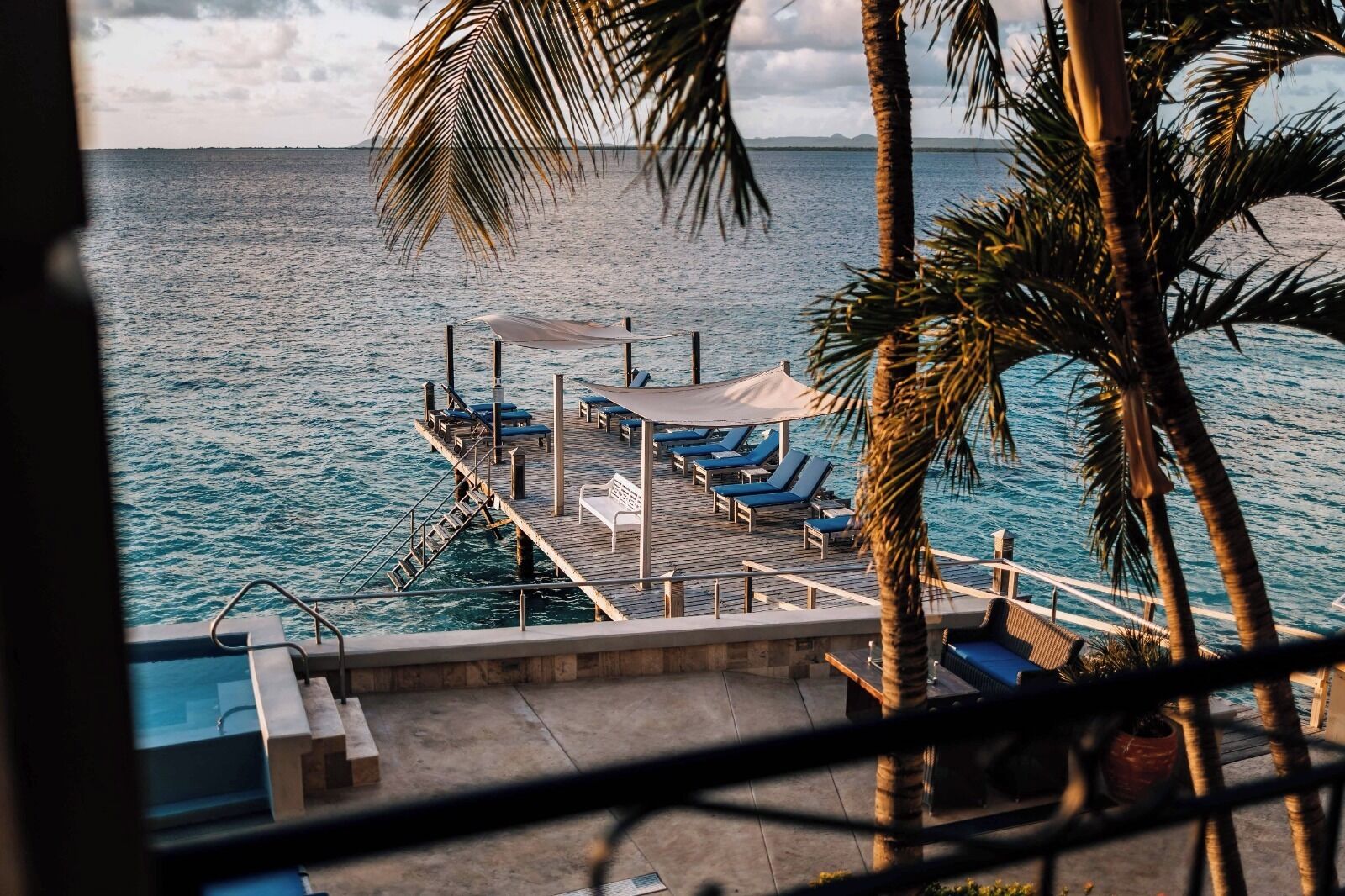 A pier with lounge chairs and a canopy, overlooking a calm sea with palm trees.