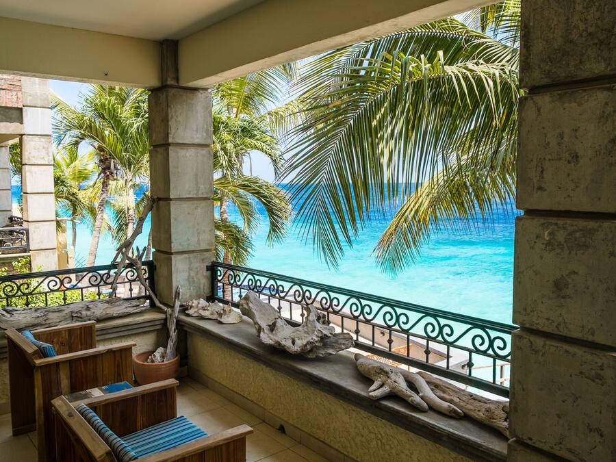 A balcony with a view of a turquoise ocean and palm trees, wooden furniture, and decorative elements.