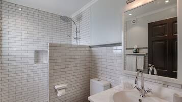 Ein Badezimmer mit weißen Fliesen im U-Bahn-Stil, einer weißen Toilette, einer Dusche mit Glastür, einem weißen Waschbecken mit Spiegel und einer dunklen Holztür.
