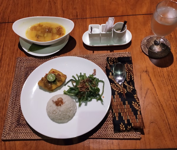 Een maaltijd met rijst, groene bonen en een currygerecht op een bord, vergezeld van een kom soep en een glas ijsthee op een houten tafel.