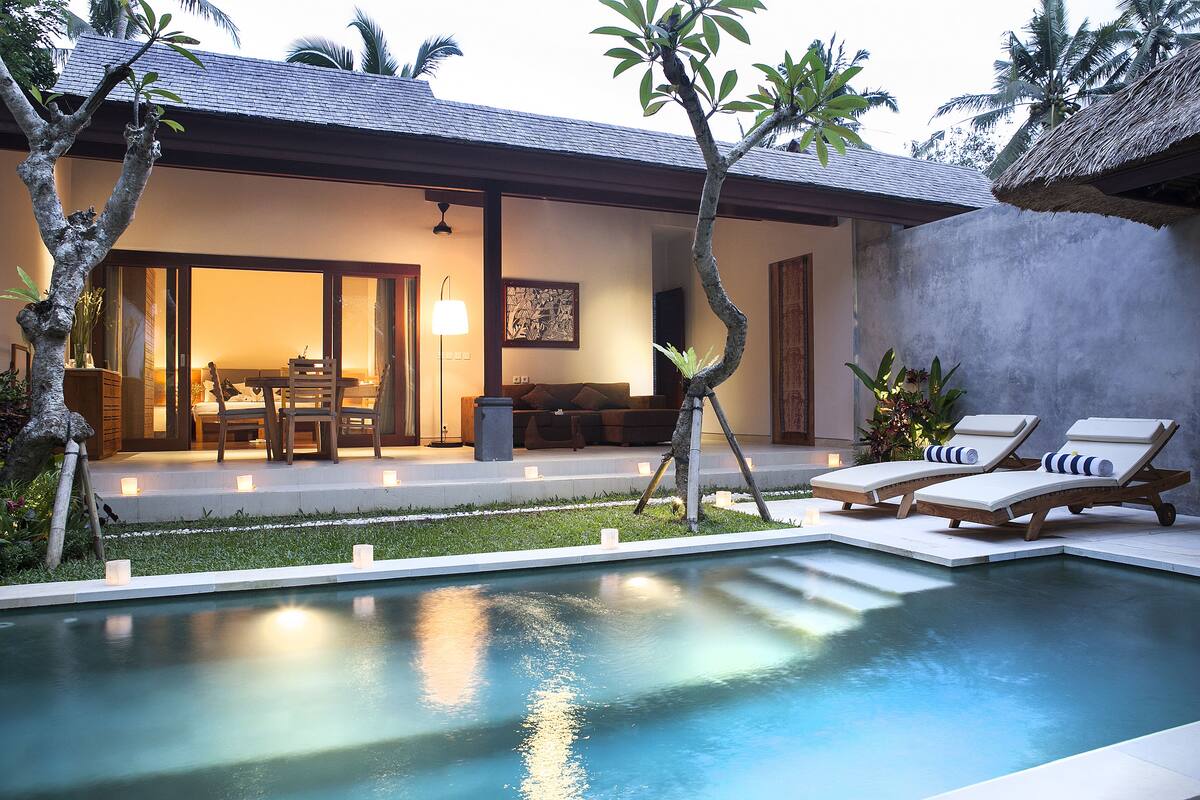 Un área junto a la piscina con sillones, una zona de comedor con mesa y sillas, y una estructura con techo de paja.