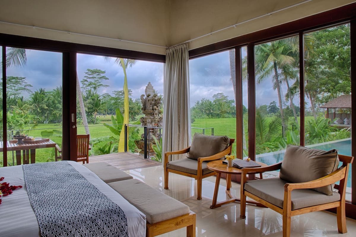 Un dormitorio con una cama grande, dos sillas, una mesita y vistas a un jardín exuberante con palmeras.