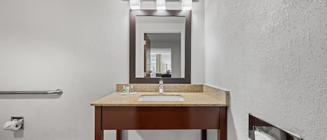 A bathroom with a wooden vanity, a sink, a mirror with lights, a towel dispenser, and a handrail.
