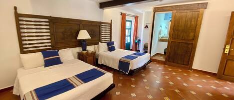 Habitación de hotel con dos camas, cabecera de madera, mesita de noche con lámpara y baño visible a través de una puerta abierta.
