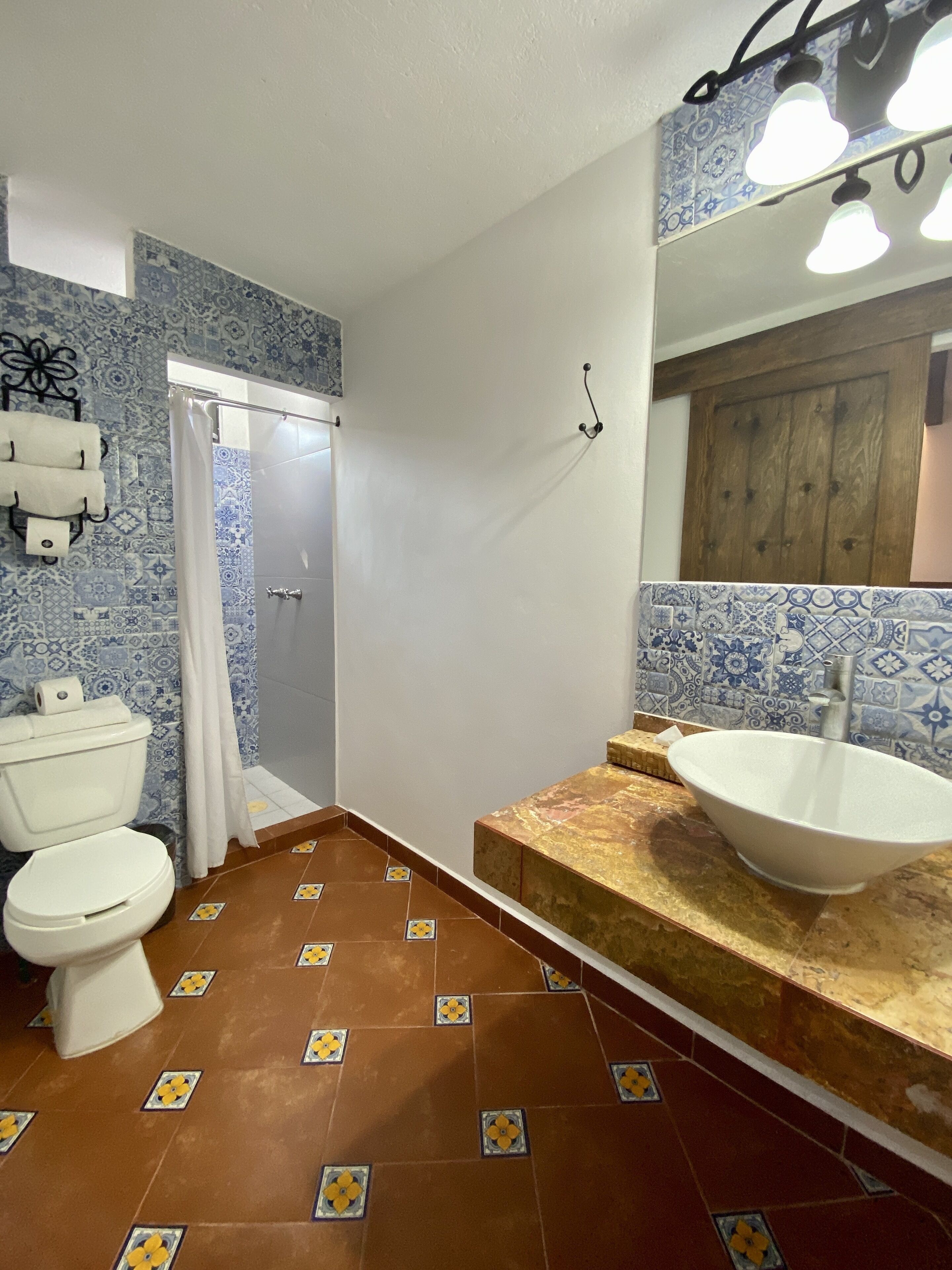 Un baño con inodoro blanco, cortina de ducha, lavamanos con encimera de mármol y azulejos estampados en paredes y suelo.