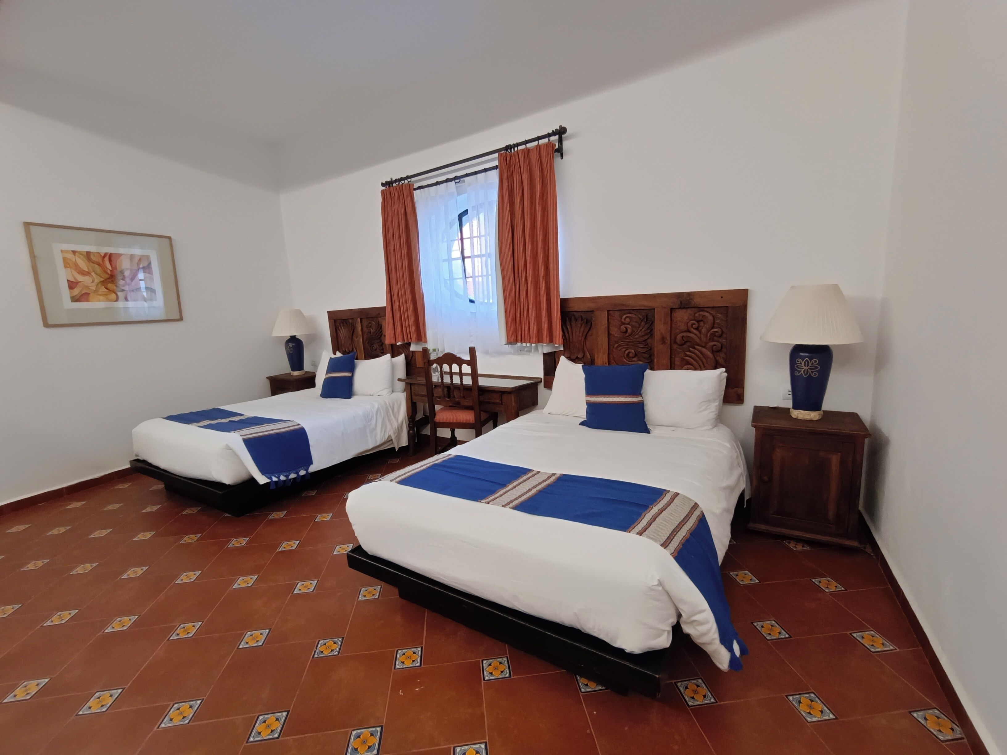 A hotel room with two beds, a wooden headboard, a chair, a table, and a window with curtains.