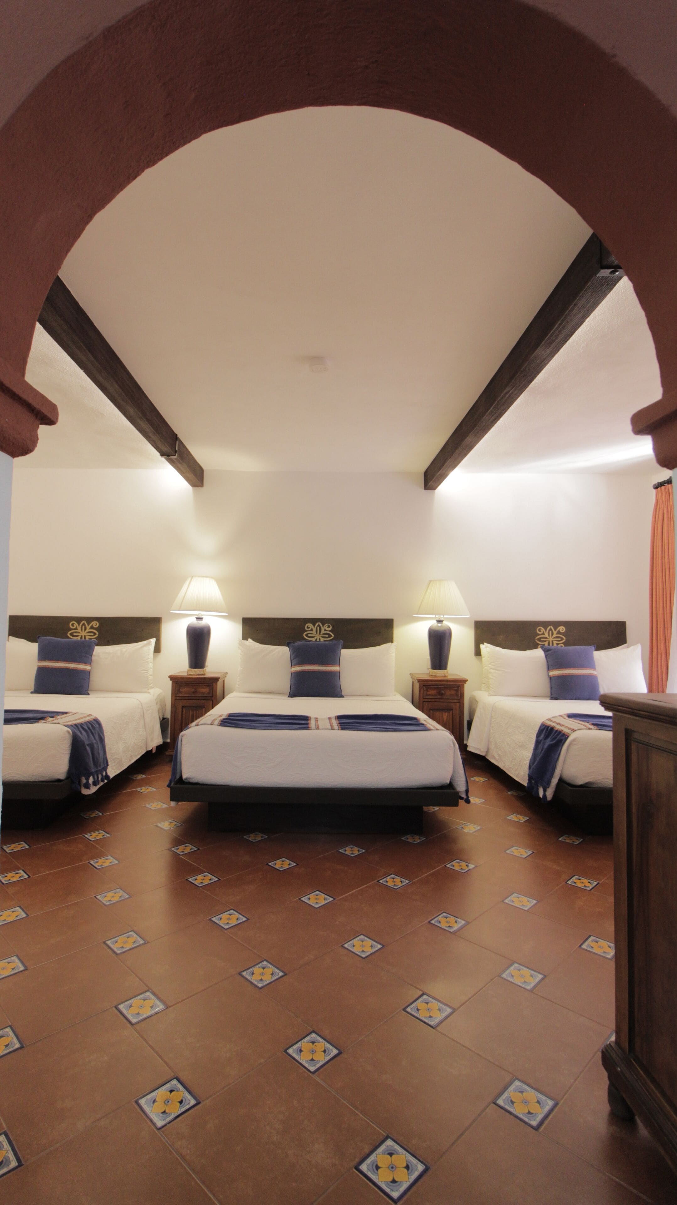 Een hotelkamer met een groot bed, twee kleinere bedden en een nachtkastje met lampen. De kamer heeft een betegelde vloer en een houten plafond.