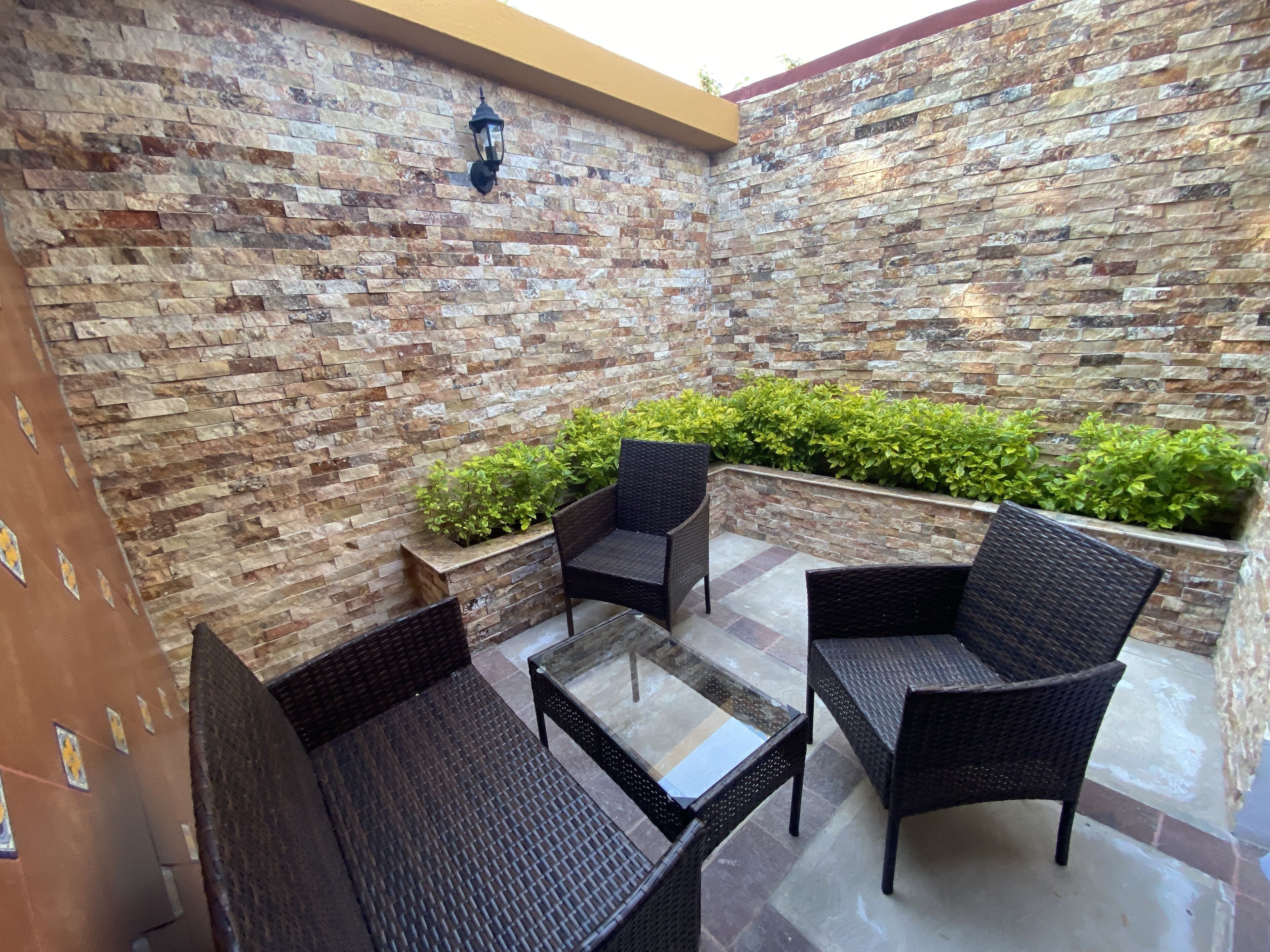 An outdoor seating area with wicker furniture and a glass-top table, surrounded by a stone wall and greenery.