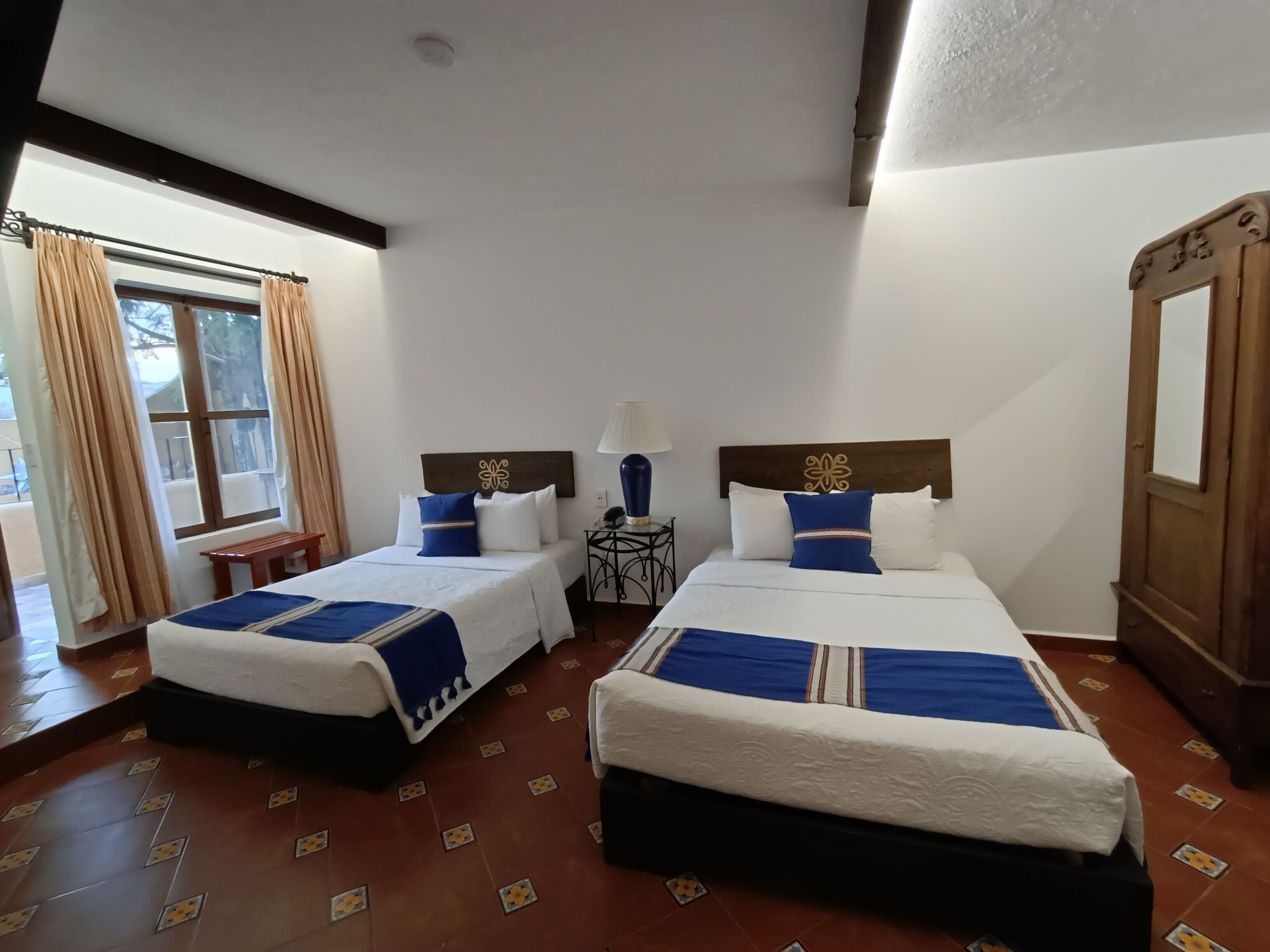 A hotel room with two beds, a wooden wardrobe, a small table, and a window with curtains.