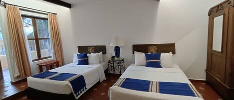 A hotel room with two beds, a wooden wardrobe, a small table, and a window with curtains.