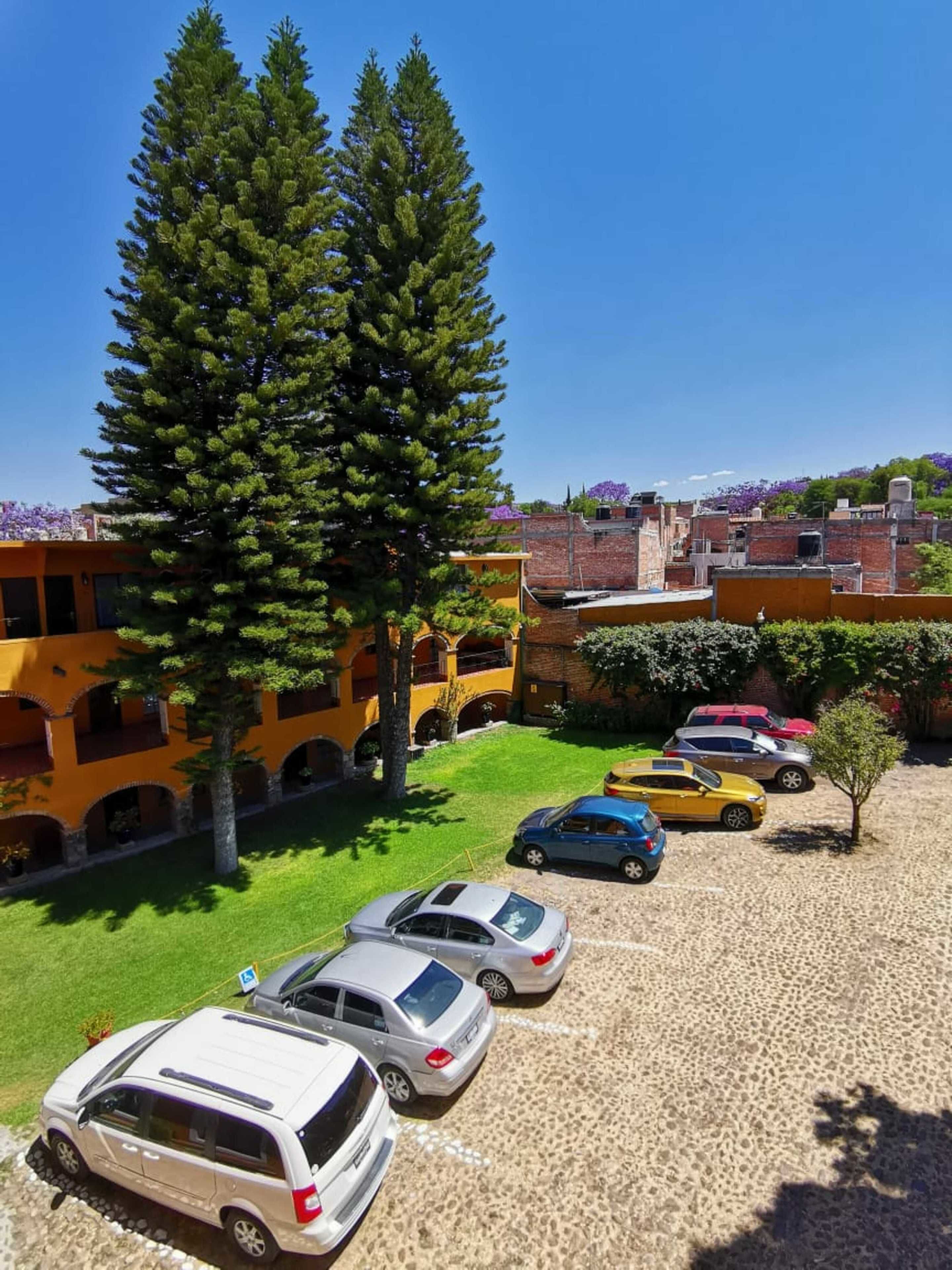 A parking area with several cars, a large tree, and a building with yellow walls and balconies.