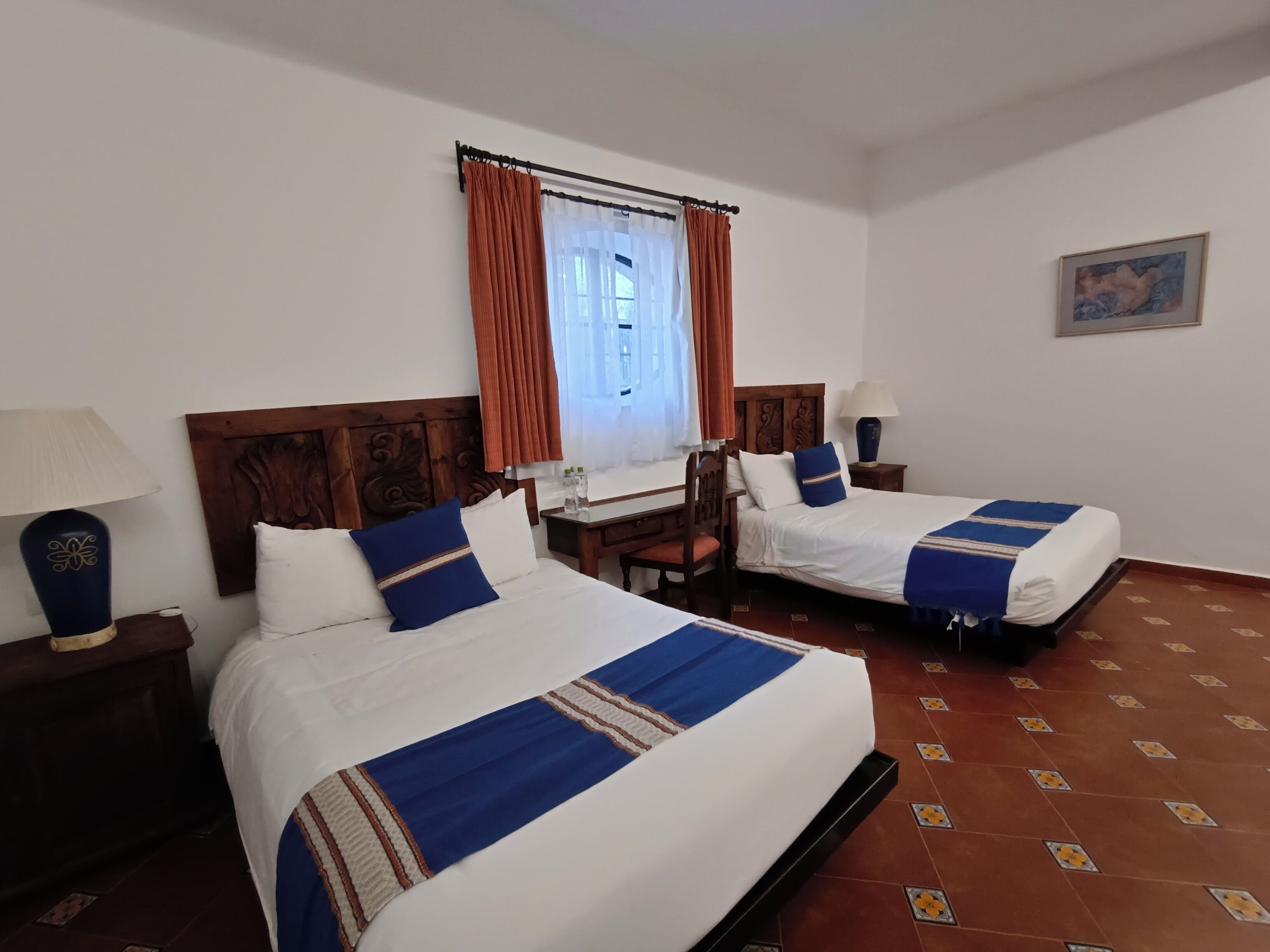 Habitación de hotel con dos camas, cabecero de madera, escritorio, silla, lámpara y un cuadro en la pared.