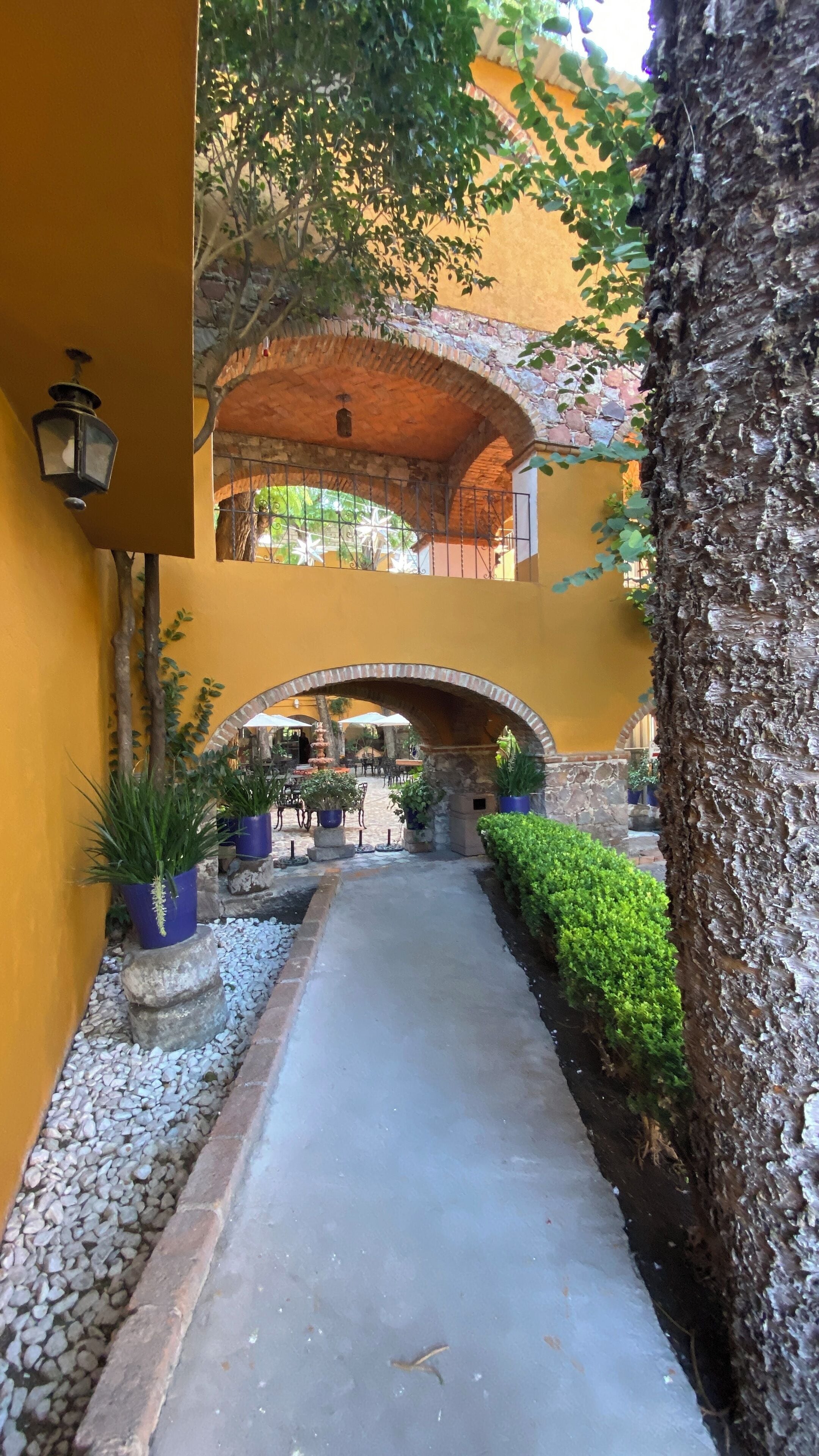 An outdoor corridor with a stone archway, potted plants, and a pathway leading to a seating area.