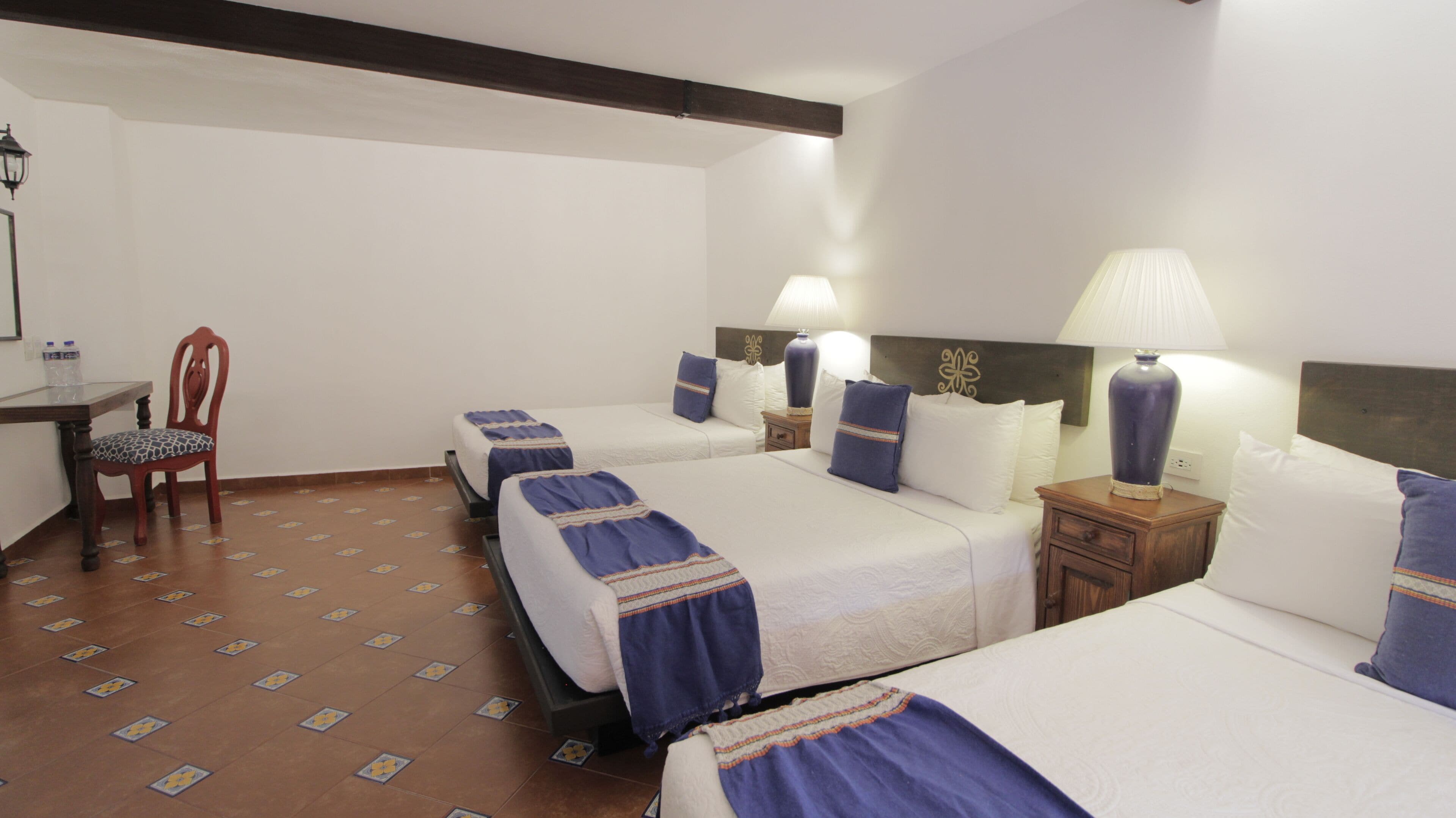 Une chambre d’hôtel avec quatre lits simples, une chaise en bois, une petite table et une lampe.