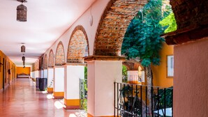 Un couloir avec des arches en briques, un sol carrelé et une rambarde ornée d’un motif végétal.