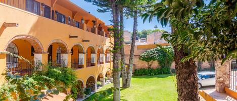 A courtyard with a yellow building featuring arched windows and balconies, surrounded by greenery and trees.