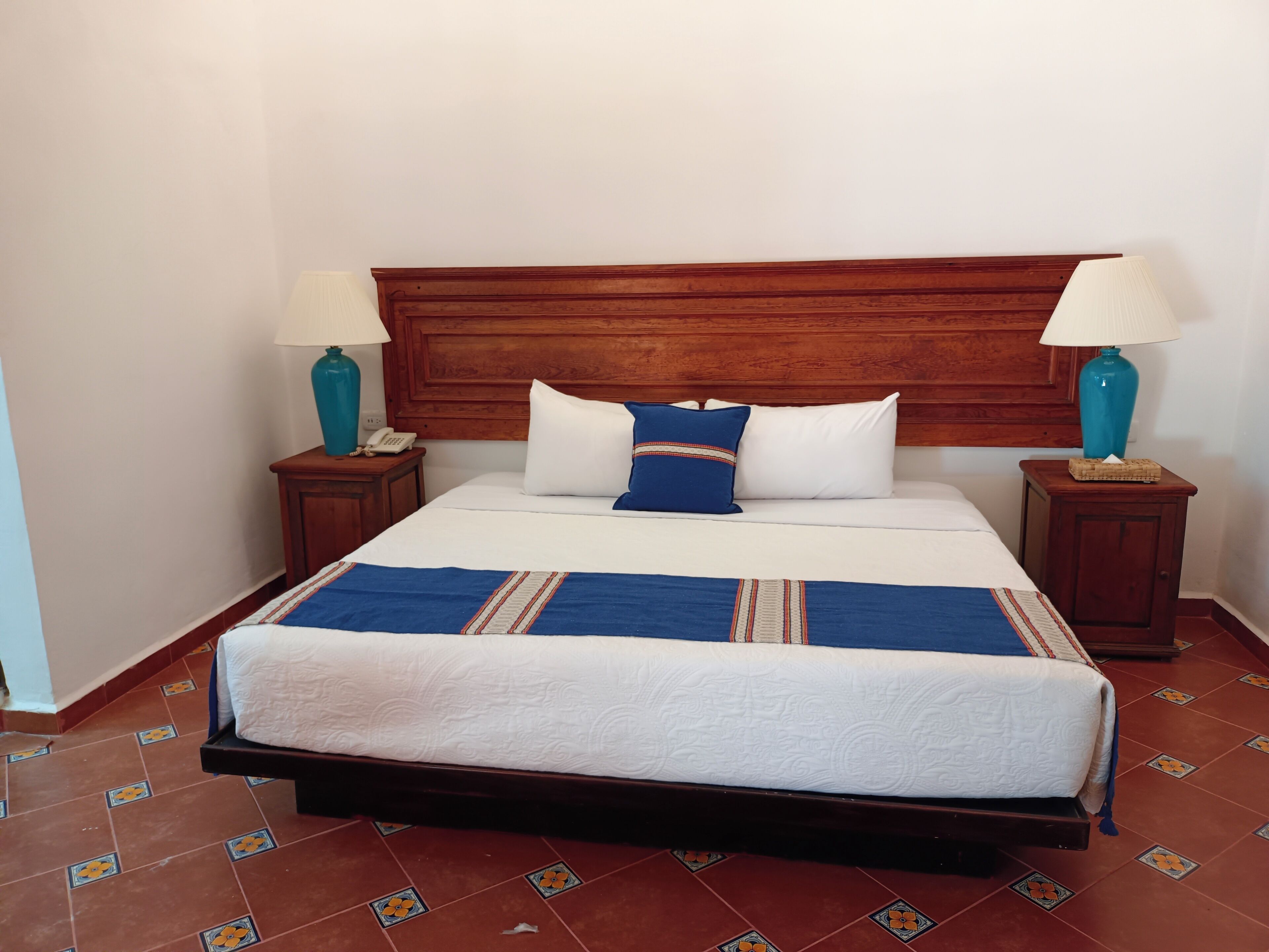 A neatly made bed with a wooden headboard, two bedside tables with lamps, and patterned floor tiles.